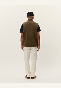 Brown fleece vest with high collar, worn over a black t-shirt, paired with cream pants and brown shoes, viewed from the back.