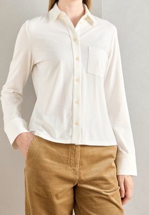 Kobieta w białej koszuli na guziki z kieszenią na piersi i beżowych spodniach sztruksowych, jedną ręką w kieszeni, stojąca na tle szarej ściany.