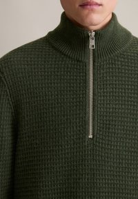 Dunkelgrüner Pullover mit hohem Kragen und halbem Reißverschluss. Strukturierter Stoff mit einem Gittermuster und gerippten Akzenten.
