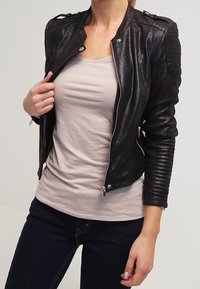 Kobieta w czarnej skórzanej kurtce, nosząca jasnoróżową koszulę i ciemne dżinsy, trzymająca klapę kurtki jedną ręką.