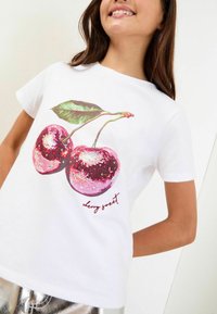 Mladý člověk v bílém tričku se dvěma lesklými, flitrovými třešněmi a zelenými listy, s nápisem "cherry sweet" pod designem.