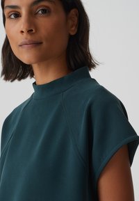 Haut vert foncé à col haut avec des manches courtes, en tissu doux et avec des détails de couture minimalistes. Conçu pour un ajustement décontracté avec une texture lisse.