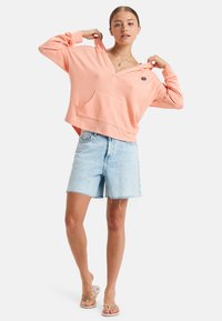 Licht perzikkleurige hoodie met een voorzak, relaxte pasvorm en een zonnenembleem. Combinatie met vervaagde denimshorts en slippers.