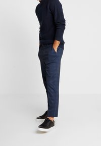 Dark blue knit sweater with texture on upper part, paired with tailored dark blue trousers and black sneakers with white soles.