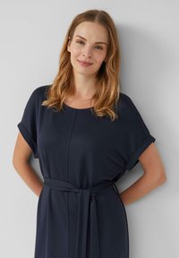 Dunkelblaues Kleid mit lockerer Passform, umgeschlagenen Ärmeln und einem Gürtel, der in der Taille gebunden ist. Weicher Stoff mit subtiler Textur und rundem Ausschnitt.
