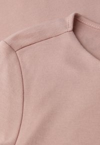 Zacht roze stof met gedetailleerde stiksels langs de schoudernaad en de halslijn van een kledingstuk.