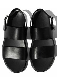 Crne kožne sandale s dva široka remena koja prelaze preko stopala, remenom oko gležnja s kopčom i ravnim potplatom. Glatka tekstura, minimalistički dizajn.