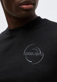 Κοντινό πλάνο ενός ατόμου που φοράει μαύρο πουκάμισο με γκρίζο λογότυπο "Mizuno" και κυκλικό σχέδιο στο στήθος.