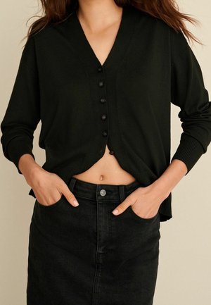 Žena v černém kardiganu na knoflíky a černé denimové sukni s vysokým pasem s rukama v kapsách, odhalující břicho.