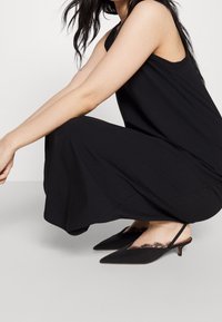 Schwarzes ärmelloses Kleid aus weichem Stoff, kombiniert mit schwarzen Slingback-Schuhen mit spitzer Zehe und Spitzenverzierungen am Rand.