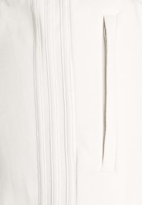 Valkoinen kangas, jossa on pystysuorat poimutukset ja sivutasku. Tekstuurin väitetään olevan sileä. Suunnittelu korostaa puhtaita linjoja ja minimalistista tyyliä.