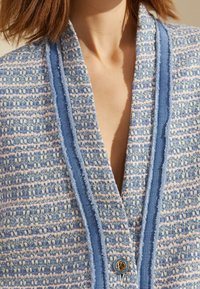 Blå och rosa texturerad kofta med djup V-ringning, med ribbat avslut och guldknaffte. Stickad tyg med intrikata mönster.