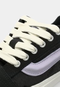 Svart sneakers med hvite snørebånd, med en stripe i lilla semsket skinn. Materialet ser glatt ut med en teksturert finish rundt snorlåsene.