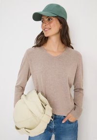 Pull beige à manches longues avec un col en V, associ�é à un denim bleu clair. Une casquette verte et un sac beige clair sont inclus dans la scène.