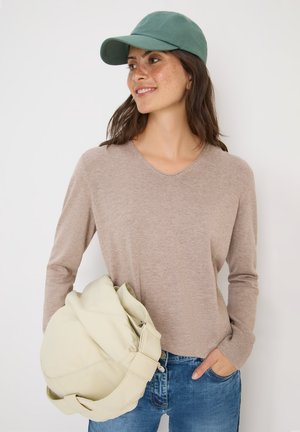 Beige Langarmpullover mit V-Ausschnitt, kombiniert mit hellblauen Jeans. Eine grüne Mütze und eine hellbeige Tasche sind ebenfalls in der Szene enthalten.