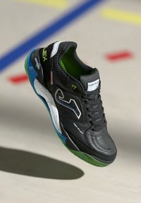 Bottines de sport noires avec une tige en cuir texturé, des accents verts, des détails bleus et blancs, et une semelle en caoutchouc conçue pour une utilisation en intérieur.