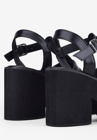 Sandalias de plataforma negras con un tacón grueso, correas de cuero suave y detalles de hebilla ajustable en el tobillo para un ajuste seguro.