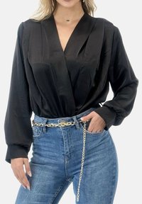 Sort bluse med lange ærmer og V-hals i blød tekstur, kombineret med blå højtaljede jeans og et guldkædebælte som detalje.