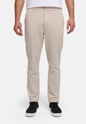 Mand iført beige teksturerede bukser og sorte afslappede sneakers, stående mod en ensfarvet hvid baggrund.