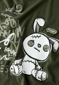 Cartoon-Hase mit gestickten Nähten und Knopfaugen, Sprechblase "MEINE REGELN" und Graffiti-Text vor einem dunkelgrünen Stoffhintergrund.