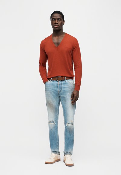 Homme noir debout, portant un pull à col en V orange rouille, un jean bleu clair déchiré, une ceinture marron et des baskets blanches, sur un fond blanc uni.