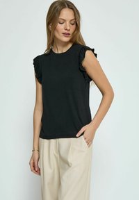 Haut noir à manches courtes avec des accents de volants sur les épaules. Tissu lisse, coupe classique, associé à un pantalon plissé beige clair.