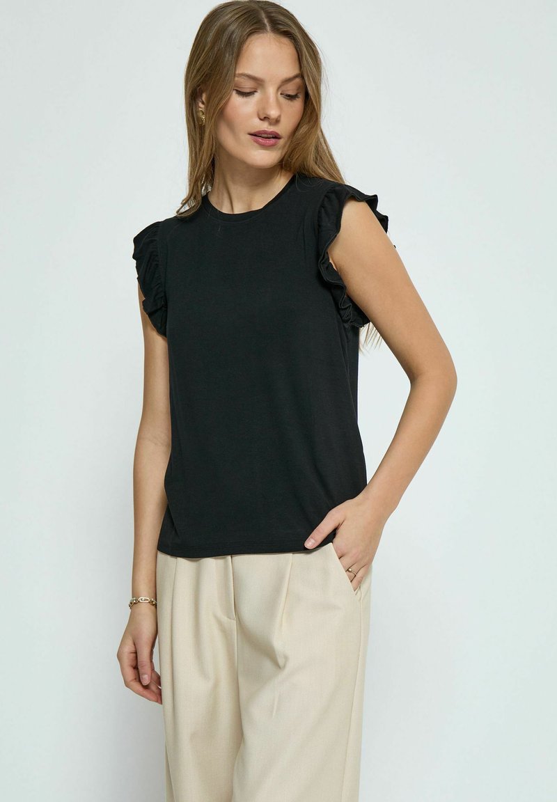 Haut noir à manches courtes avec des accents de volants sur les épaules. Tissu lisse, coupe classique, associé à un pantalon plissé beige clair.