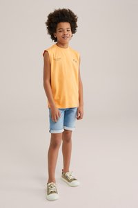 Oranžové tielko bez rukávov s potlačou palmy; svetlomodré džínsy s krátkymi nohavicami a ohrnutými manžetami; béžové tenisky s bielymi podrážkami a šnúrkami.