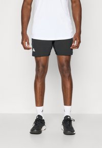 Sorte atletiske shorts med et tekstureret design og hvidt Adidas-logo, parret med sorte løbesko og hvide ankelstrømper. Hvid t-shirt.