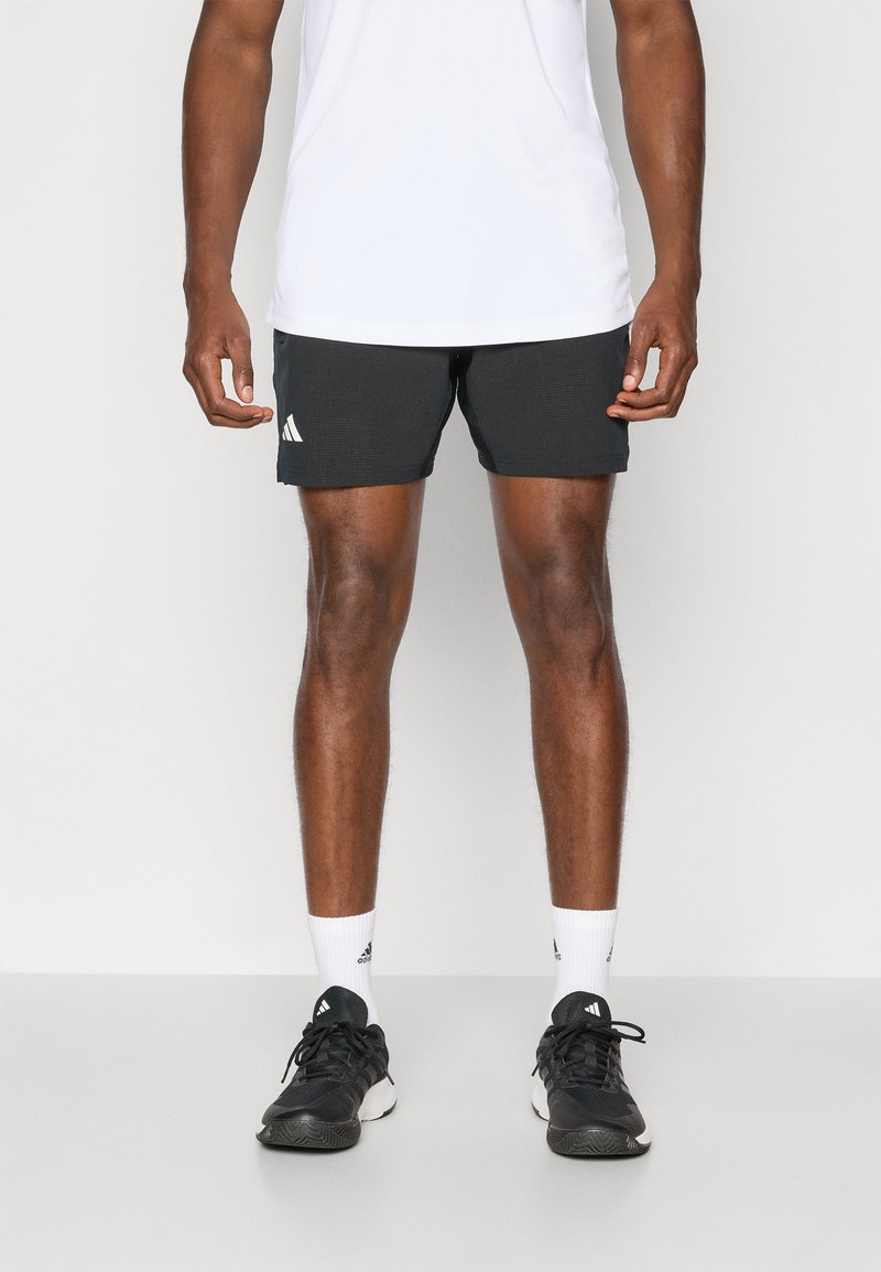 Sorte atletiske shorts med et tekstureret design og hvidt Adidas-logo, parret med sorte løbesko og hvide ankelstrømper. Hvid t-shirt.