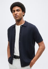 Άνδρας με κοντά σγουρά μαλλιά που φοράει μπλε σκούρο κοντομάνικο πουκάμισο με κουμπιά πάνω από λευκό μπλουζάκι, ανοιχτόχρωμο παντελόνι και μαύρη ζώνη, με τα χέρια στις τσέπες.