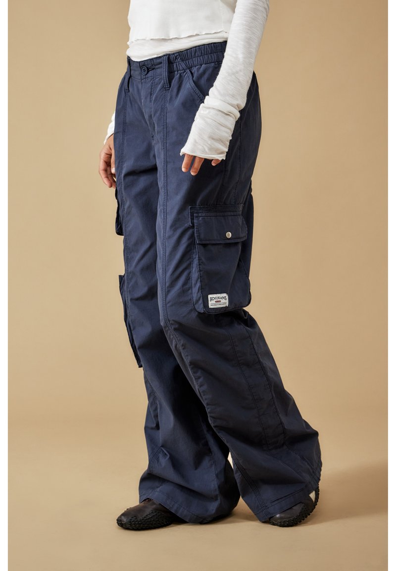 Person klädd i marinblå cargobyxor med sidofickor och en vit långärmad skjorta, stående mot en beige bakgrund.