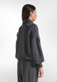 Pullover grigio oversize con collo alto, maniche plissettate e polsini elastici. La parte posteriore presenta un piccolo zip rosa come accento. Texture morbida.