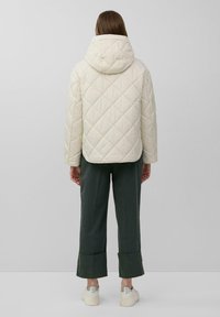 Gepolsterte cremefarbene Jacke mit Kapuze; zeigt ein Rautenmuster. Kombiniert mit dunkelgrünen, knöchelfreien Hosen und weißen Turnschuhen. Einfaches Design.