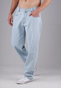 Person, der hellblaue Jeans und weiße Sneakers trägt, steht mit einer Hand in der Tasche vor einem grauen Hintergrund.