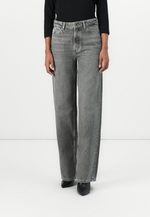 Personne portant un jean droit gris délavé taille haute, des bottes noires à bout pointu et un haut noir à manches longues sur un fond blanc.