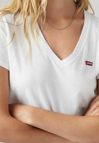 Camiseta de algodón blanca con un diseño de cuello en V. Presenta un pequeño logo rojo de Levi's en el lado izquierdo del pecho. Mangas cortas y un ajuste relajado.