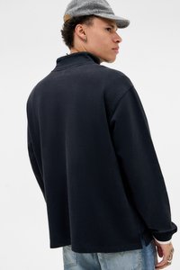 Ung mand iført en sort sweatshirt, gråternet kasket, lyseblå jeans og sølvørering, der kigger til højre mod en hvid baggrund.