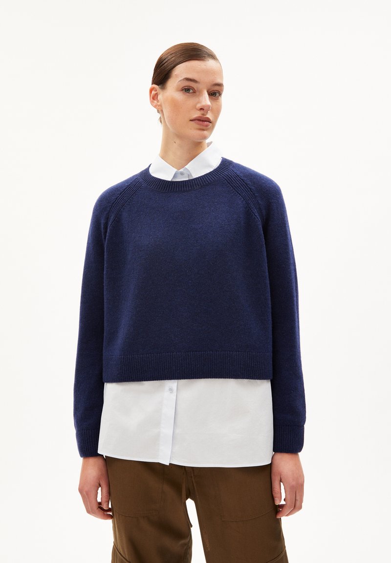 Marineblauer, kurz geschnittener Pullover aus weichem Strick mit gerippten Bündchen und Saum, getragen über einem weißen Hemd mit Kragen. Einfaches, schlankes Design.