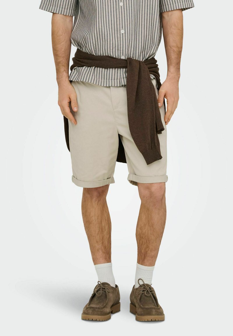 Homme portant un short beige retroussé, des chaussettes blanches, des chaussures en daim marron, une chemise à manches courtes à motifs et un pull marron noué autour de la taille.