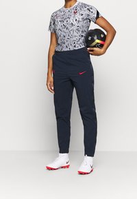 Conjunto de ropa deportiva Nike que incluye una camiseta de manga corta de color gris con estampado, pantalones marinos y zapatillas blancas con acentos rojos, sosteniendo un balón de fútbol negro.