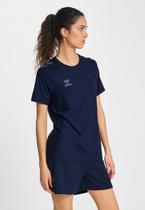 Donna con capelli ricci raccolti in una coda di cavallo, che indossa una maglietta sportiva blu navy e pantaloncini, in piedi di profilo su sfondo bianco.