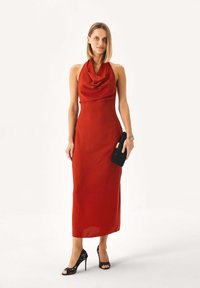 Nainen, jolla on punainen hihaton midi-mittainen halter-kaulainen mekko, mustat pitsikorkokengät, musta clutch-laukku kädessään, seisoo valkoista taustaa vasten.