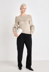 Beige off-shoulder tröja med ribbade detaljer, tillsammans med svarta vida byxor. Modellen håller en vit strukturerad handväska. Svarta spetsiga skor.