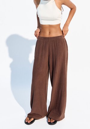 Persoon die losse bruine broek en een wit kort tanktop draagt, en de tailleband aanpast, staand tegen een effen lichte achtergrond.