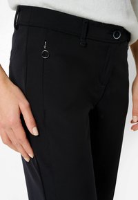 Pantalon noir sur mesure avec une texture lisse, coupe slim, et une poche zippée ornée d'un anneau métallique au niveau de la taille.