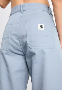 Pantalons en coton bleu clair avec une taille haute, des poches latérales et deux poches arrière arborant un patch logo carré. Texture lisse, coupe décontractée.
