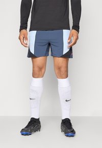 Manlig idrottare iklädd en svart långärmad tröja, blå och svarta shorts, vita knähöga Nike-strumpor och svarta fotbollsskor med blå dobbar.