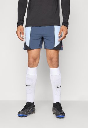 Male athlete wearing black long-sleeve shirt, blue and black shorts, white knee-high Nike socks, and black soccer cleats with blue studs.