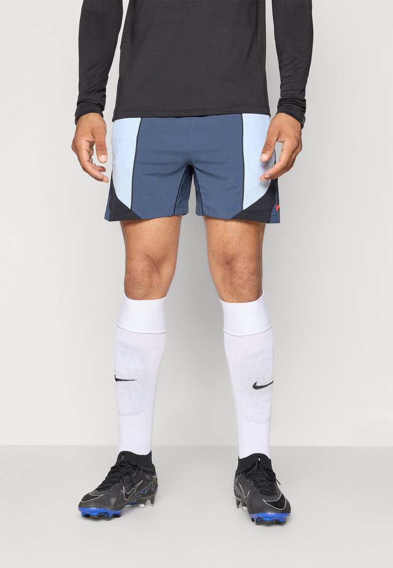 Manlig idrottare iklädd en svart långärmad tröja, blå och svarta shorts, vita knähöga Nike-strumpor och svarta fotbollsskor med blå dobbar.